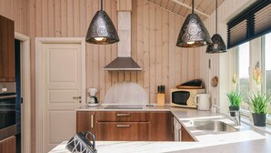 A modern kitchen with wooden panel walls, a central island with a sink, a microwave, and a coffee maker. There are three pendant lights above the island and two potted plants by the window. - 4 Star Holiday Home in Hemmet (Hemmet)