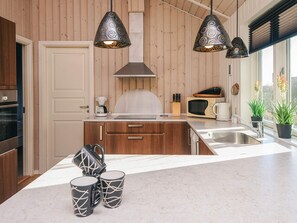 A modern kitchen with wooden panel walls, a central island with a sink, a microwave, and a coffee maker. There are three pendant lights above the island and two potted plants by the window. - 4 Star Holiday Home in Hemmet (Hemmet)