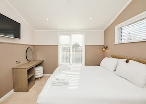 A modern bedroom with a bed, a bedside table, a mirror, a wall-mounted TV, and a window with blinds.