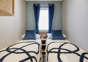 A room with two beds, a window with curtains, a bedside table with a lamp, and a wall-mounted shelf.