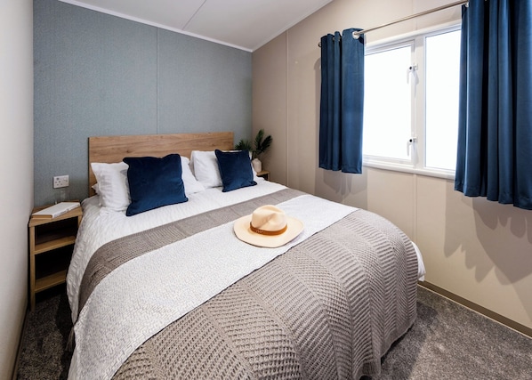A neatly made bed with a white and gray blanket, a beige hat, and blue pillows. - 3 Bed Holiday Home PF (Peterborough)