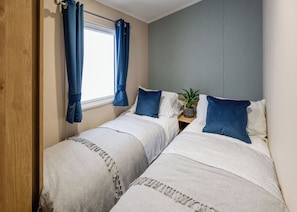 Una habitación con dos camas, una ventana con cortinas, una planta sobre una mesita de noche y una puerta de madera.