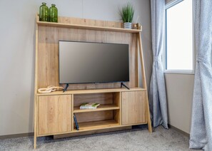 A wooden TV stand with a flat-screen TV, a shelf with green bottles, a potted plant, and a window with curtains. - 3 Bed Holiday Home PF (Peterborough)