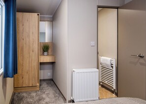 A bedroom with a wooden wardrobe, a small bedside table with a plant, a radiator, and a door leading to another room. - 3 Bed Holiday Home PF (Peterborough)