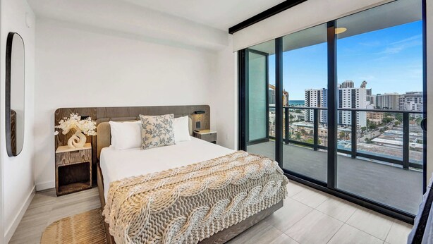 Une chambre moderne avec un grand lit, une table de chevet, un miroir et une vue sur la ville grâce à de grandes fenêtres.