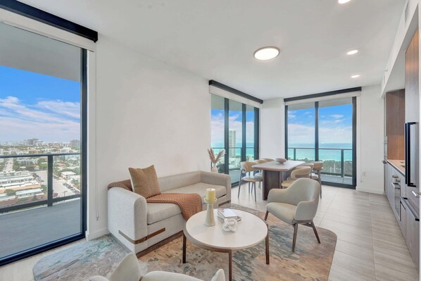 A modern living room with a sofa, coffee table, and dining area, large windows with a city view, and a balcony.