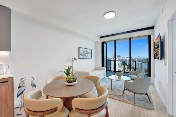 A modern living room with a round dining table, white sofas, and a large window with a city view.