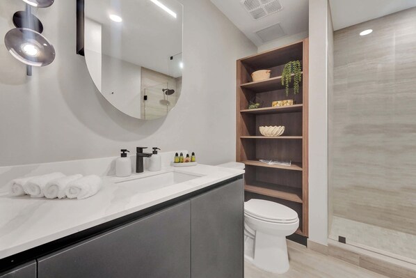 Un baño moderno con un lavamanos, espejo y retrete blancos. Hay toallas, un dispensador de jabón y una estantería con adornos.