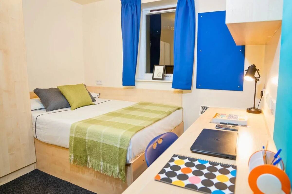 A small, well-lit room with a bed, a desk, and a chair. There is a window with curtains, a lamp on the desk, and a notebook with a colorful cover.
