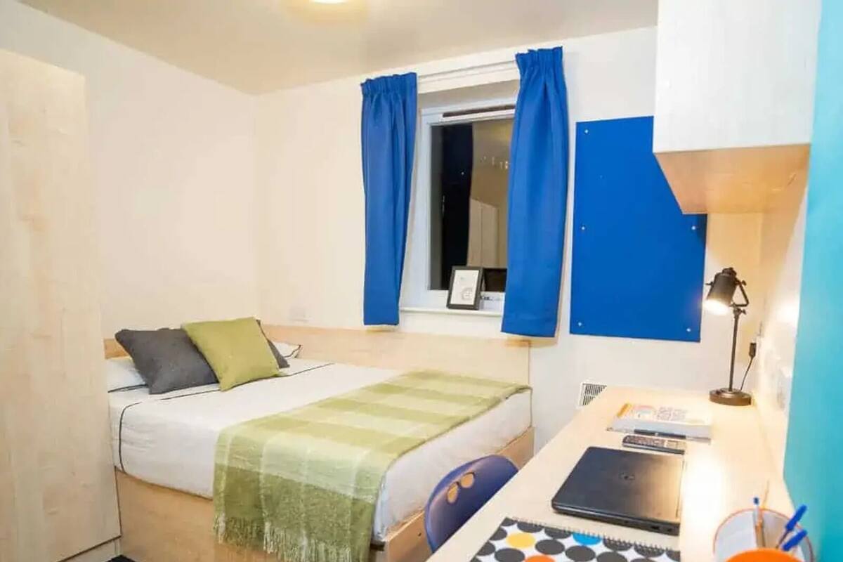 A compact hotel room with a bed, a desk with a laptop, a notebook, and a desk lamp.