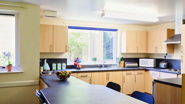 A modern kitchen with wooden cabinets, a black countertop, a window with blue blinds, and a bowl of fruit on the counter. - For Students Only Queen's Hospital Close near Birmingham City Centre (Birmingham)
