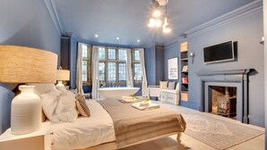 A bedroom with a bed, bedside tables, a fireplace, a window, and a television. - Luxurious 4 bedroom apartment @Pickwell Manor on North Devon Coast near Croyde (Georgeham)