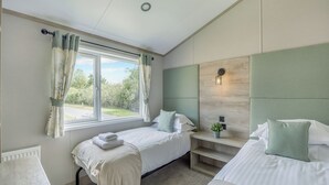 A bedroom with two beds, a window with curtains, a bedside table with a lamp, and a view of greenery outside.