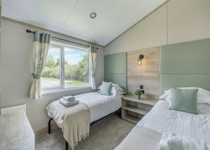 A bedroom with two beds, a window with curtains, a bedside table with a lamp, and a view of greenery outside. - 2 bedroom accommodation in Woodhall Spa (Woodhall Spa)