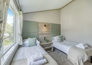 A hotel room with two beds, a window with curtains, a bedside table with a plant, and a wall-mounted light fixture.
