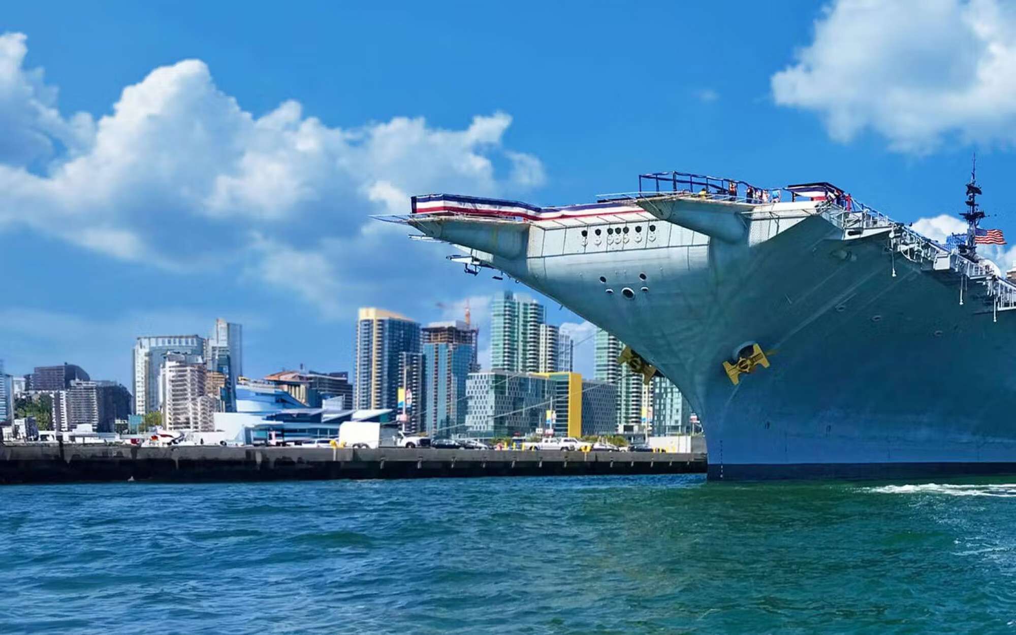 Een groot marineschip ligt aangemeerd aan een steiger, met een stadsachtergrond op de achtergrond.