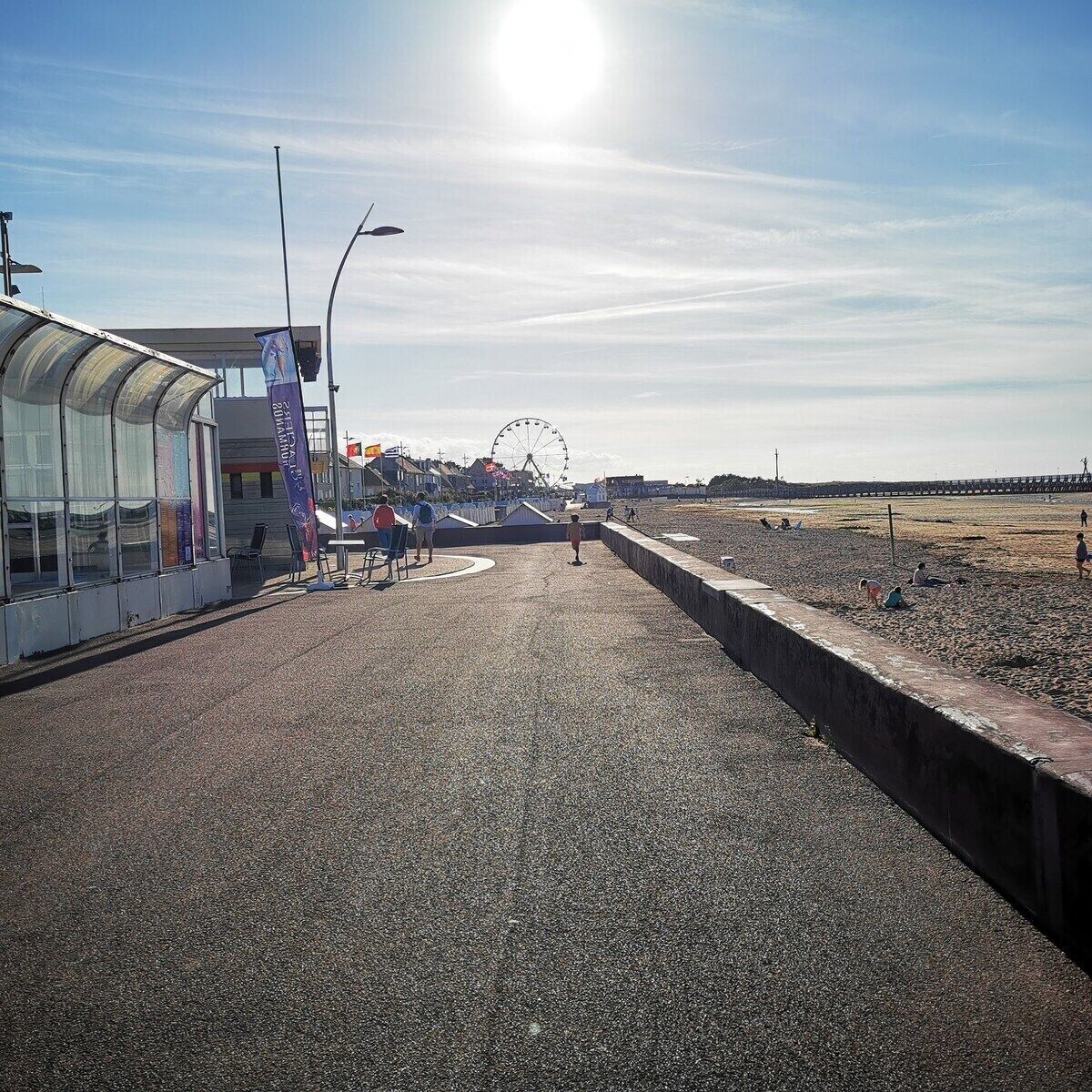 A beachside promenade with a glass building, a Ferris wheel in the background, and a clear sky.
