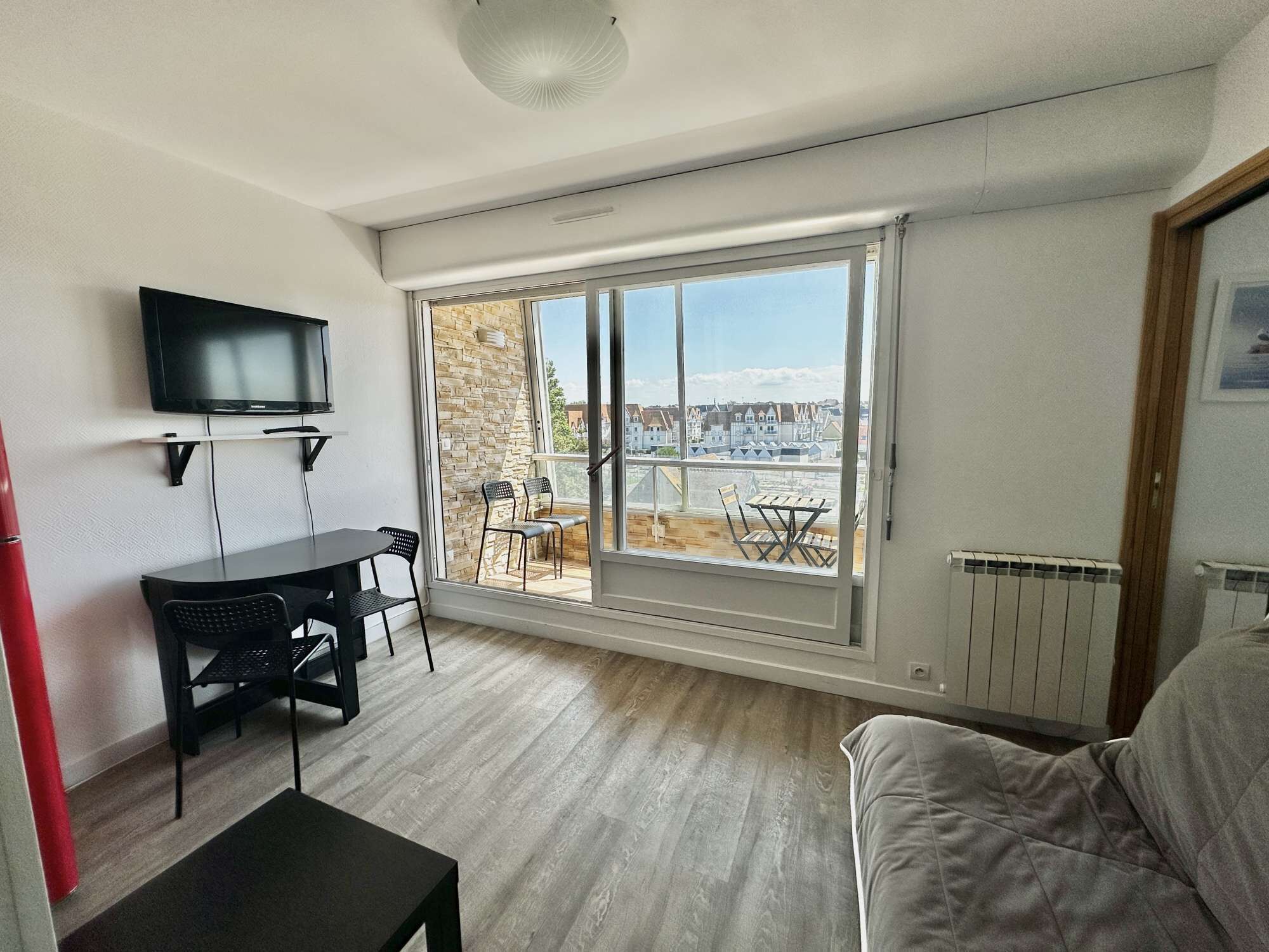 Ein Zimmer mit an der Wand befestigtem Fernseher, einem Esstisch mit Stühlen, einer Couch und einem großen Fenster mit Blick auf eine Wohngegend.