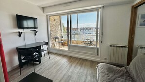 A room with a TV mounted on the wall, a dining table with chairs, a sofa, and a large window offering a view of a residential area. - Les pieds dans l'eau - south-facing loggia (Courseulles-sur-Mer)