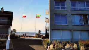 A building with a flat roof and multiple windows, located near a beach with two flags flying. - Les pieds dans l'eau - south-facing loggia (Courseulles-sur-Mer)