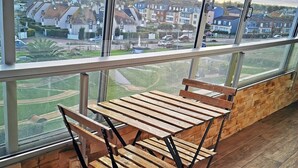 A balcony with a wooden table and chairs, large windows offering a view of a residential area. - Les pieds dans l'eau - south-facing loggia (Courseulles-sur-Mer)