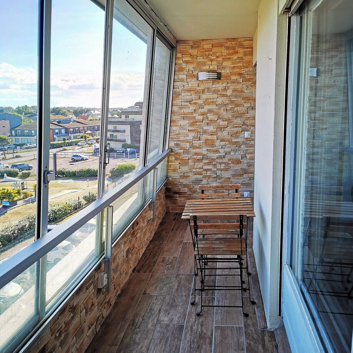 A balcony with a stone wall, a wooden table, and large windows overlooking a residential area.