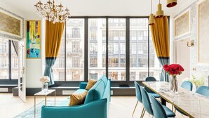 A modern living room with a large window, teal sofas, a marble dining table, and a chandelier.