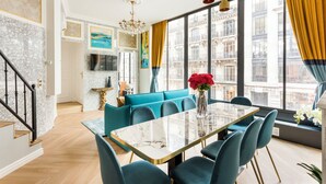 A modern living room with a marble-topped dining table, teal sofas, and a large window with yellow curtains.