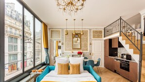 A modern hotel room with a large bed, a dining area, a kitchen, and a staircase.
