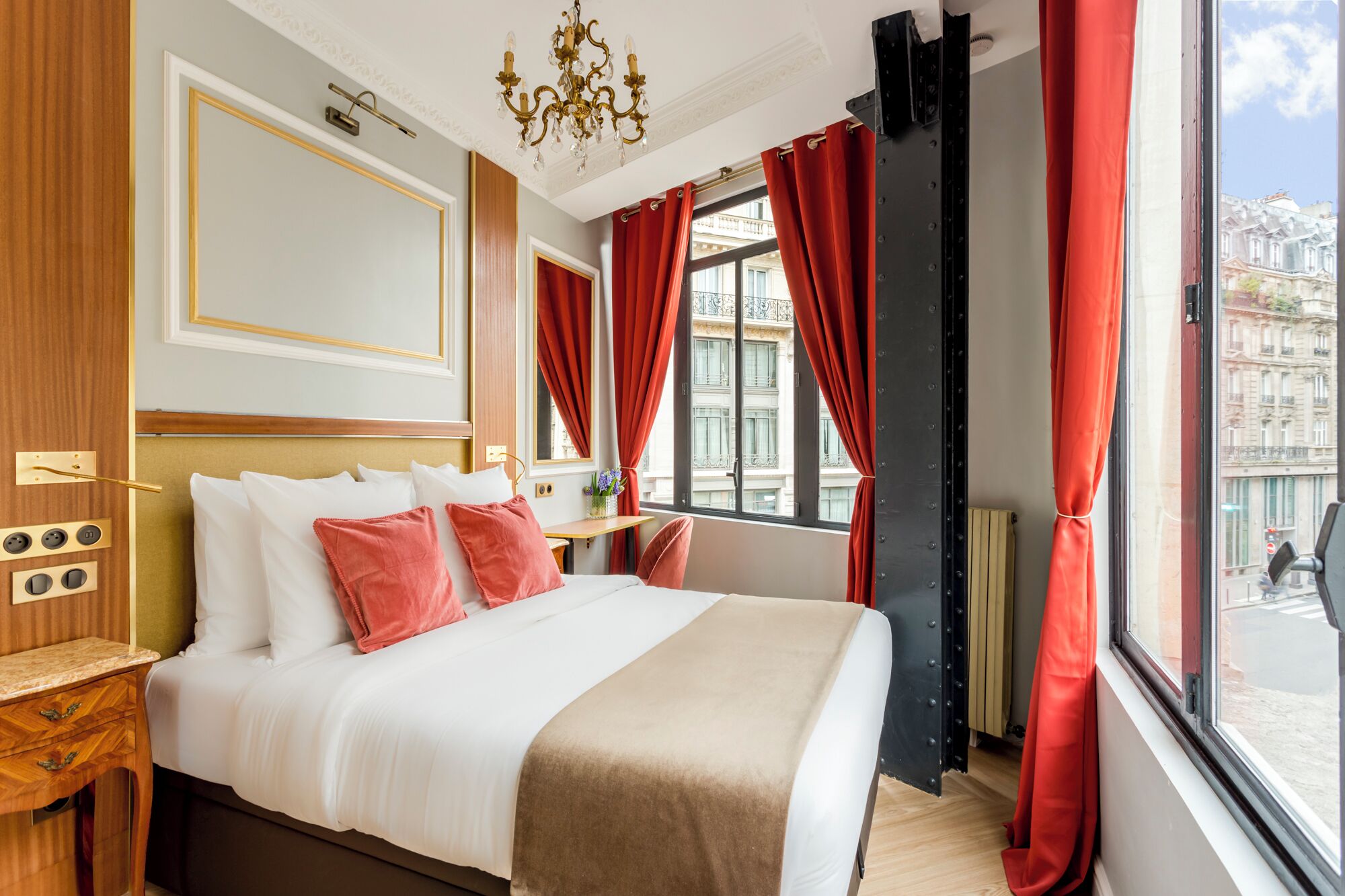 Ett hotellrum med en stor säng, röda gardiner, en kristallkrona och utsikt över byggnaderna utanför.