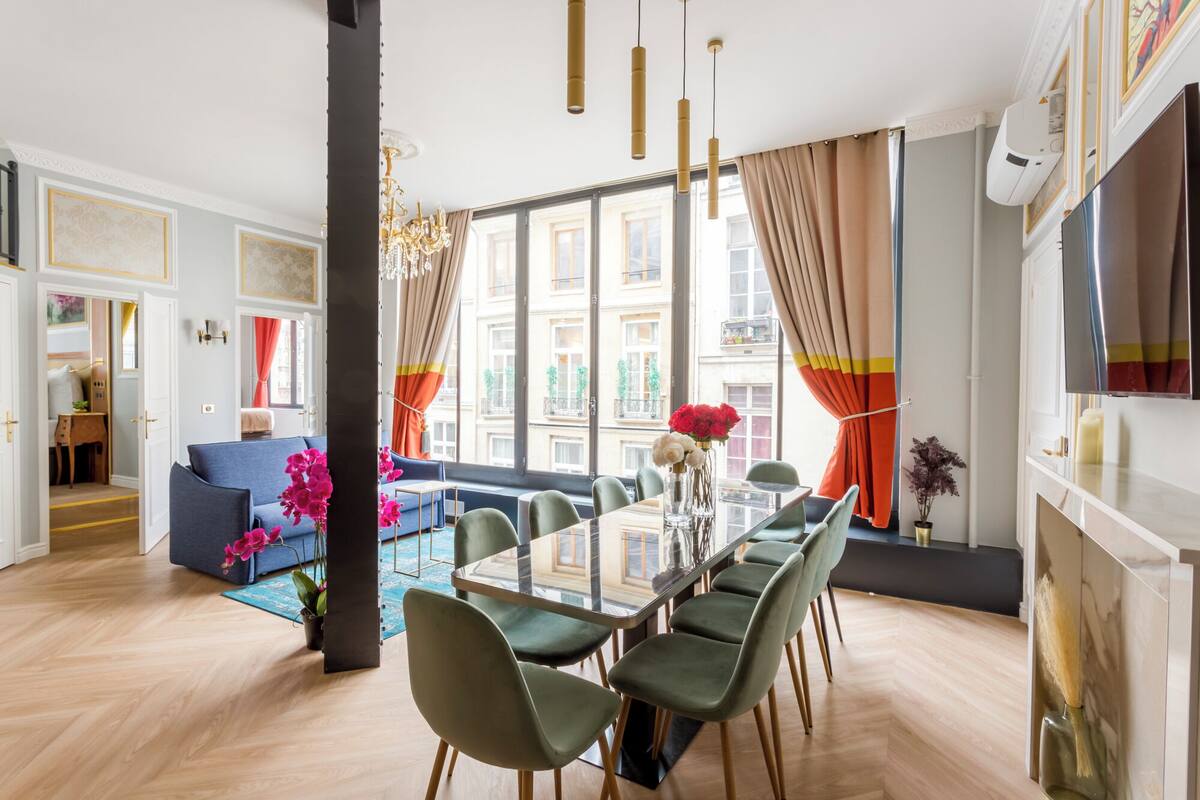 Ampla sala de estar com mesa de jantar, cadeiras e sofá. Há uma janela grande com cortinas, um lustre e uma lareira.