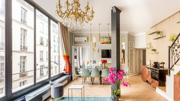 A modern living room with a large window, a chandelier, a dining area with chairs, and a kitchen with a stove.