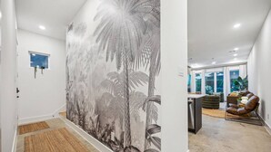 A hallway with a large wall mural of tropical foliage, a door with a small window, and a kitchen area with a dining table, chairs, and a sofa. - `Bali in Houston`/8BR/8Bath/Heated Pool/Sleeps 23 (Houston)