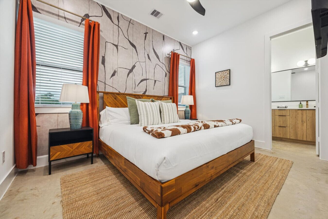 Ein modernes Schlafzimmer mit einem Holzbett, zwei Nachttischen, einem großen Fenster mit Jalousien und einem durchgehenden Türspalt sichtbaren Badezimmer.