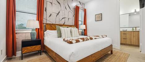 Ein modernes Schlafzimmer mit einem Holzbett, zwei Nachttischen, einem großen Fenster mit Jalousien und einem durchgehenden Türspalt sichtbaren Badezimmer.