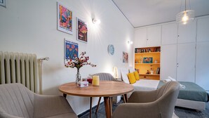 A modern room with a round wooden table, two beige chairs, a bed with green bedding, a bookshelf, and wall art. - Plaka City Studio – Walk to Syntagma by ΜΡS (Athina)