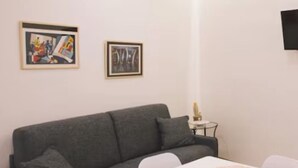 Ein Wohnzimmer mit einem grauen Sofa, einem weißen Tisch mit orangefarbener Unterlage und zwei gerahmten Kunstwerken an der Wand.