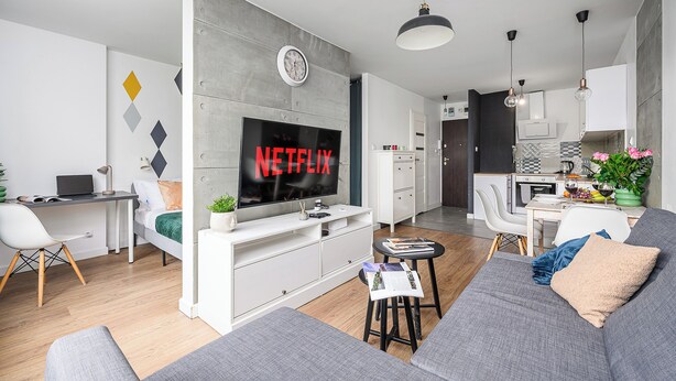 Un salon moderne doté d’un grand téléviseur à écran plat, d’un canapé gris, d’un bureau blanc et d’un coin repas avec une chaise et une table blanches.
