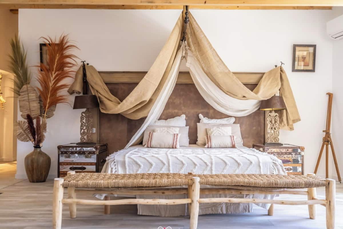 Une chambre à coucher avec un lit à baldaquin, une tête de lit en bois, des tables de chevet, un banc et des plantes décoratives.