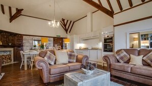 A spacious living room with a fireplace, two sofas with plaid cushions, a coffee table with pinecones, and a kitchen in the background. - Affinity@Pickwell Manor - huge space for 7 people. North Devon coast near Croyde (Georgeham)