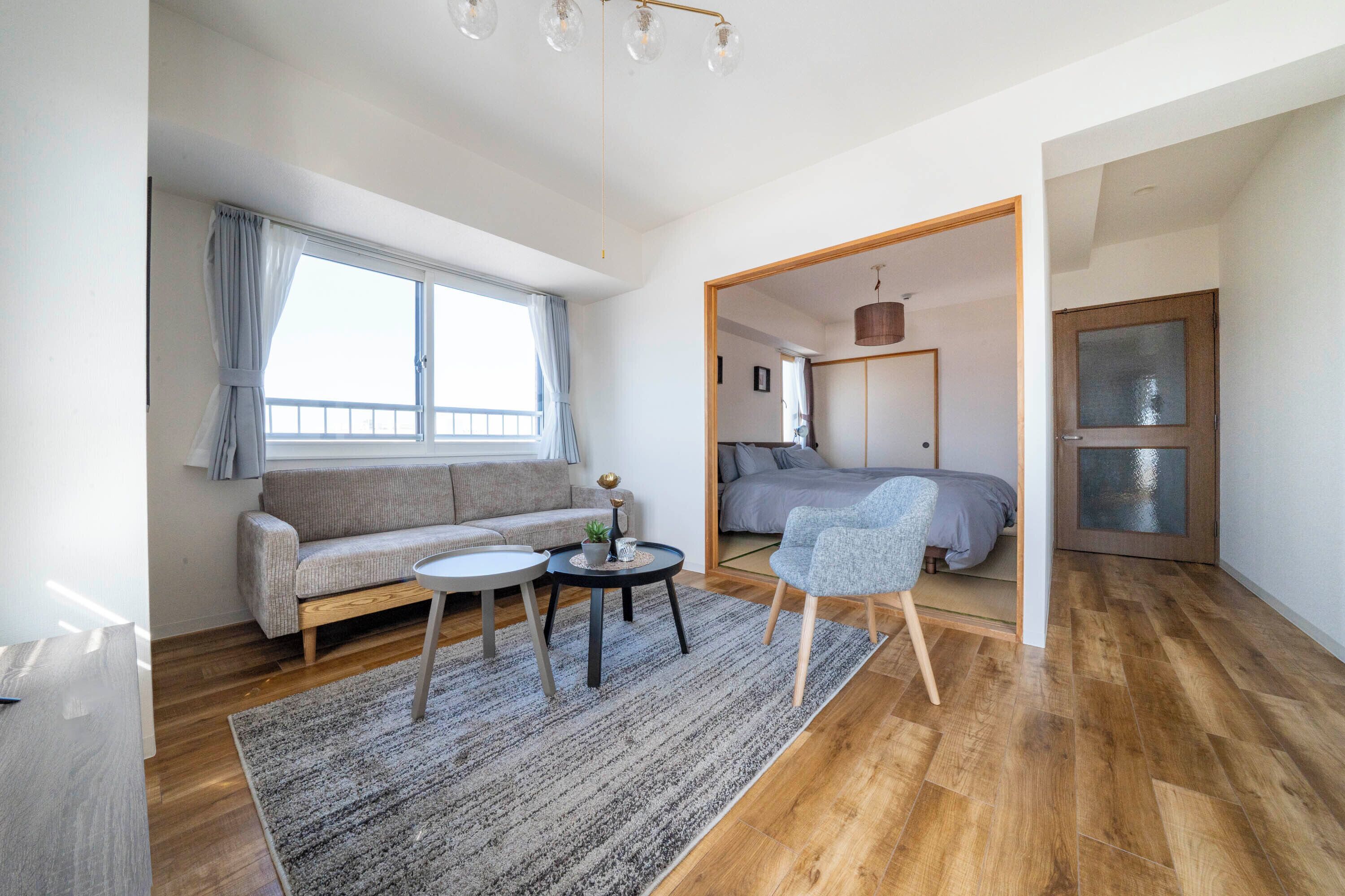 Una sala de estar moderna con un sofá, una mesa redonda, y una silla. Hay un ventanal con cortinas, una puerta que da a otra habitación, y piso de madera.