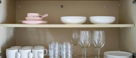 A kitchen shelf with white mugs, clear glasses, and white plates.