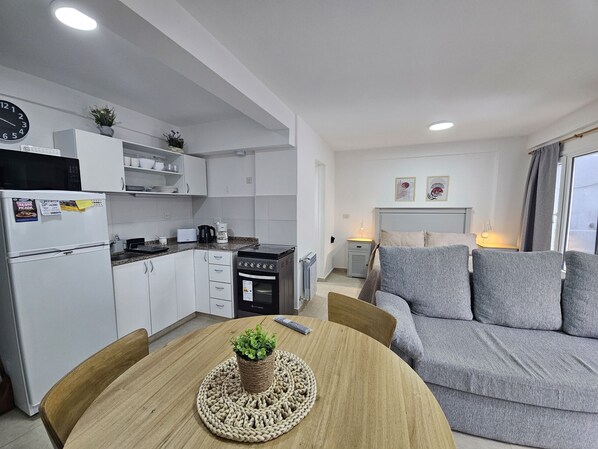 Una cocina moderna con armarios blancos, un microondas, una cafetera y un tostador. Hay una mesa de comedor con un mantel tejido y una planta en maceta. Un sofá gris se encuentra en la zona de estar, con una ventana y una obra de arte en la pared.