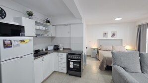 Una cocina compacta con armarios blancos, microondas, hervidor de agua y tostadora. Incluye un horno pequeño y un lavabo. La habitación tiene un sofá gris y una cama con cabecera.