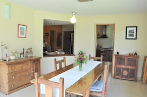 Un comedor con una mesa de madera, sillas, un jarrón con flores, un aparador y una cocina al fondo.