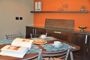 A dining area with a wooden table, chairs, and a book. There is a shelf above the table with decorative items.