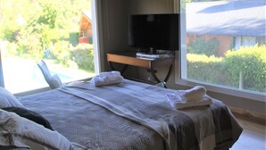 Un dormitorio con cama, televisor sobre un escritorio y vistas a una piscina y vegetación.