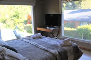 Un dormitorio con cama, televisor sobre un escritorio y vistas a una piscina y vegetación.