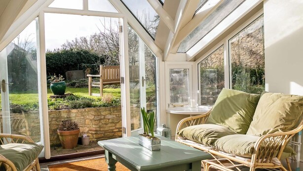 Un invernadero con techo de vidrio, mobiliario de mimbre y vistas a un jardín.