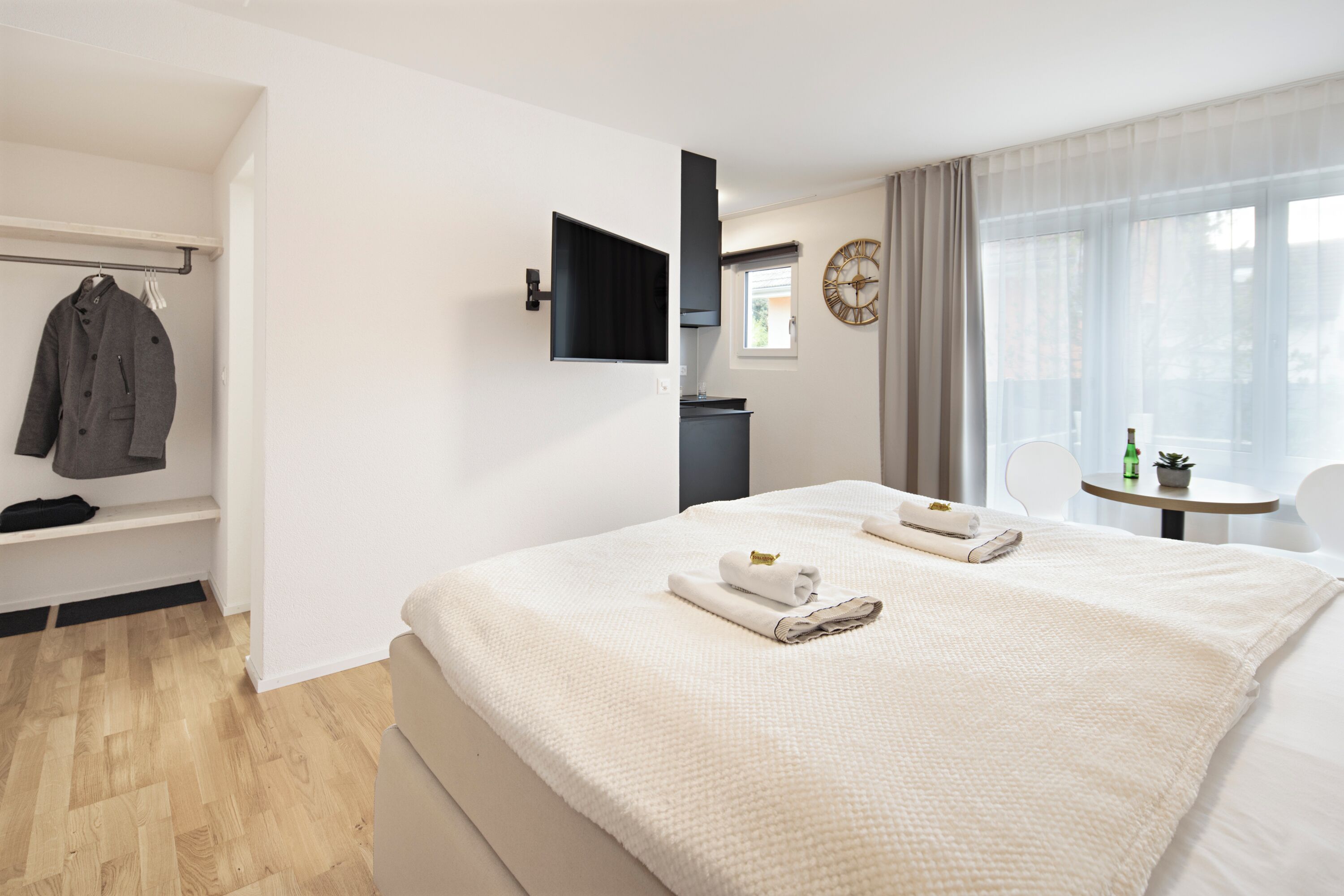 A hotel room with a large bed, a TV mounted on the wall, a small table with a plant, and a window with curtains.